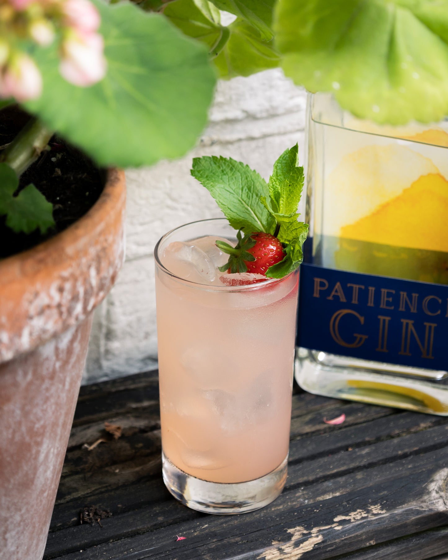 Cocktail with a strawberry and mint leaf next to a bottle of Patience Summer Gin on a wooden surface.